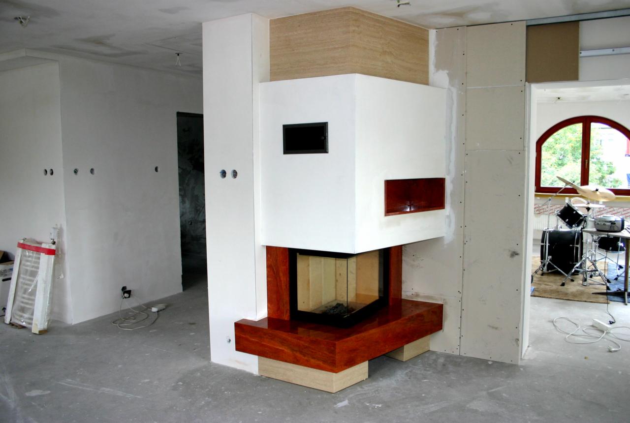 Nowoczesny kominek z drewnianą obudową w surowym wnętrzu z perkusją w tle. Widoczne płyty gipsowe i elementy wykończeniowe, sugerujące etap prac wykończeniowych.