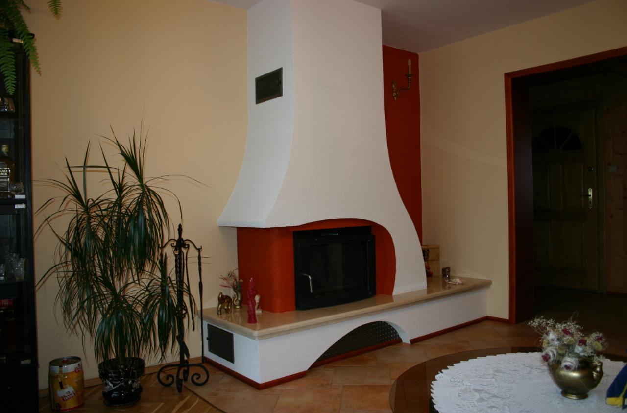 Otynkowany kominek z czarnym wkładem, obudowany płytą i pomalowany na biało, z elementami w kolorze ceglastym. Obok rośliny doniczkowe i ozdobne akcesoria.