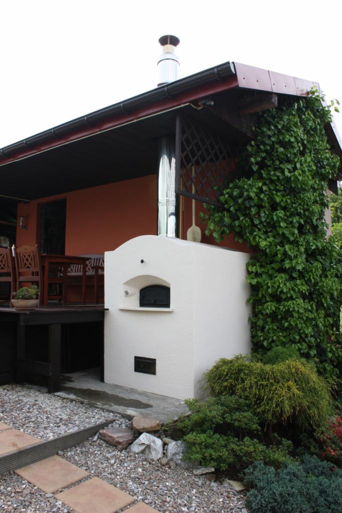 Biały piec chlebowy z metalowym kominem na zewnątrz budynku, obok drewnianego tarasu i ściany porośniętej bluszczem, z kamiennym podłożem.