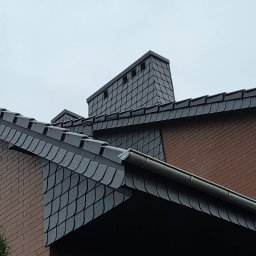 M-deal - Szary dach z łupka z kominem na tle pochmurnego nieba, widoczna ceglana ściana budynku i fragment roślinności.