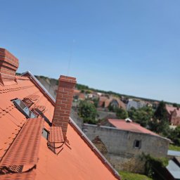 M-deal - Czerwony dach z ceglanymi kominami i oknami dachowymi w słoneczny dzień. Widok na zabudowę miejską w tle, z perspektywy z góry.