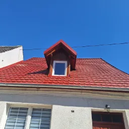 Czerwony dach z oknem dachowym i nową rynną na tle błękitnego nieba. Widoczna część budynku z oknami. Dachówka karpiówka.