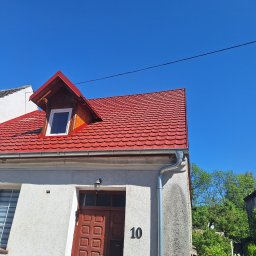 M-deal - Czerwony dach z lukarną na jasnym budynku, widoczne rynny i numer domu '10'. Błękitne niebo w tle.
