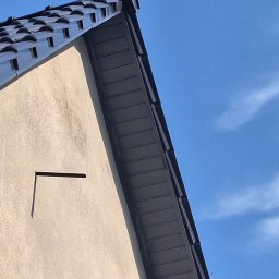 M-deal - Fragment dachu z ciemną dachówką ceramiczną i fragment ściany budynku z elementem mocowania na tle błękitnego nieba.