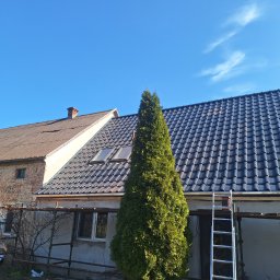 M-deal - Dom z nowym, ciemnym dachem z dachówki ceramicznej, widoczna drabina oparta o ścianę. W tle stary budynek z cegły i niebieskie niebo.