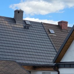 M-deal - Nowy, grafitowy dach z dachówki ceramicznej, z oknami dachowymi i kominem z cegły. Widoczny fragment dachu z gontu bitumicznego na pierwszym planie.
