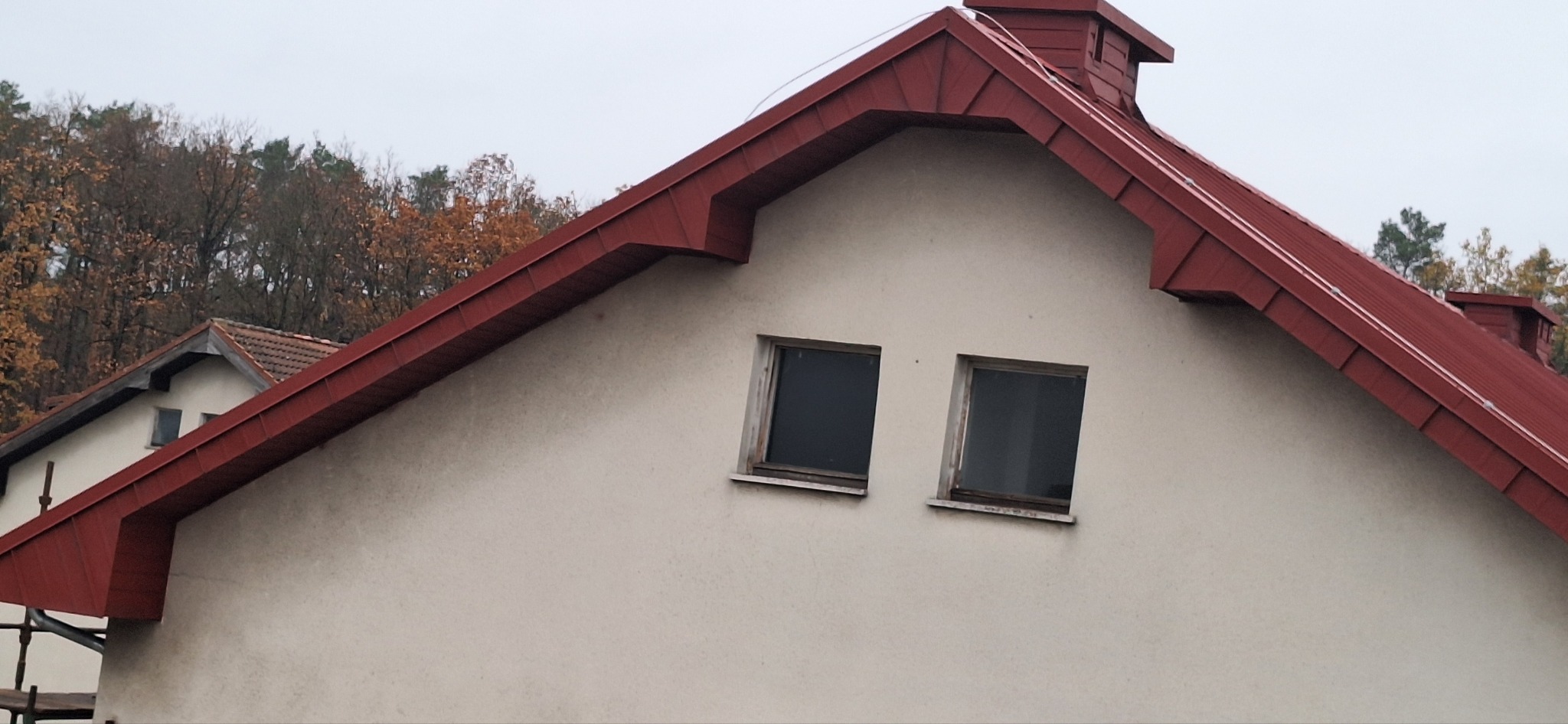 Dwuspadowy dach z czerwonej blachy na poddaszu z dwoma oknami, widoczne drzewa w tle.
