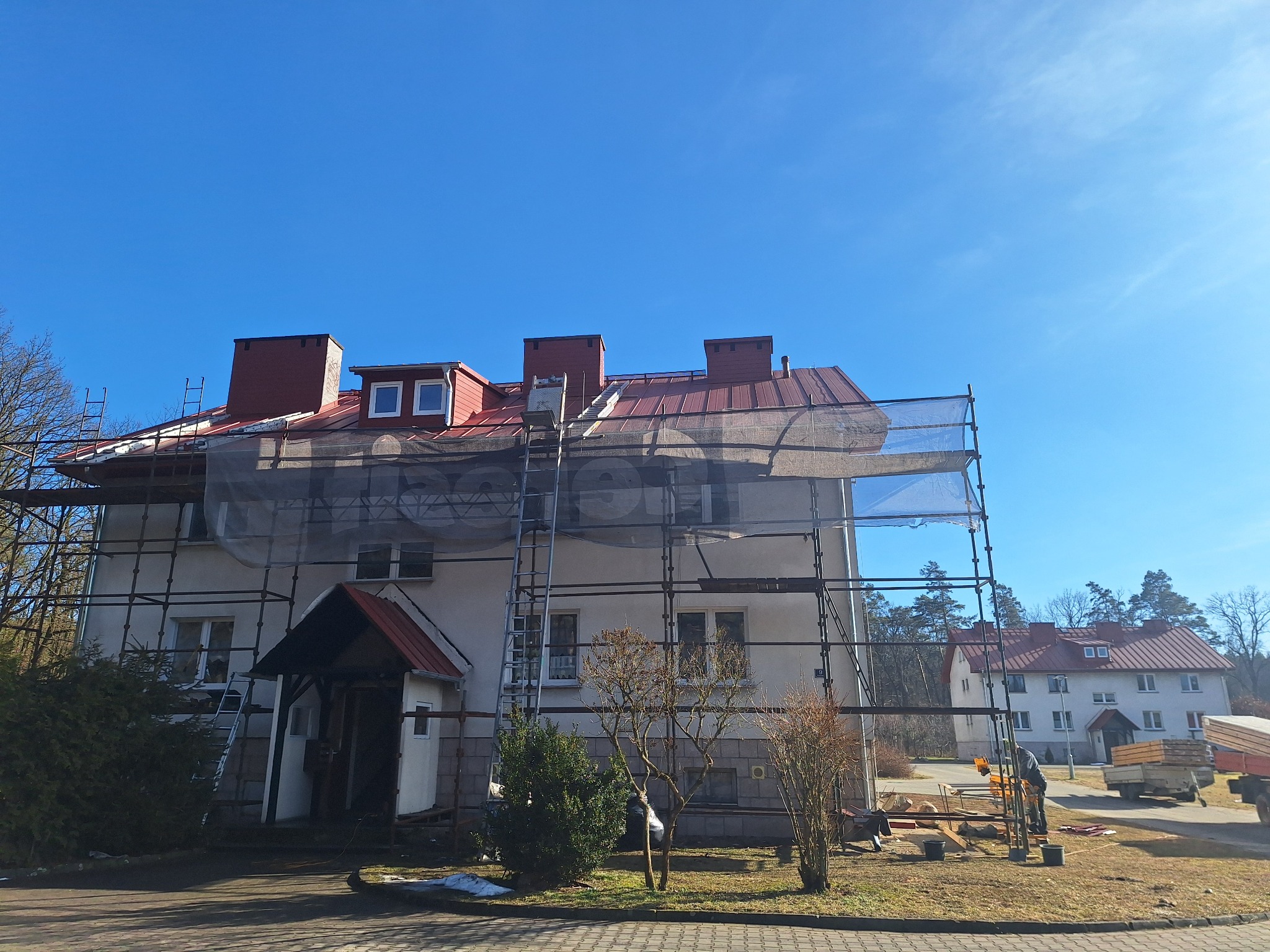 Dwupiętrowy budynek mieszkalny w trakcie prac remontowych dachu, z widocznymi rusztowaniami i siatką ochronną, czerwony dach z kominami i oknami dachowymi na tle błękitnego nieba.