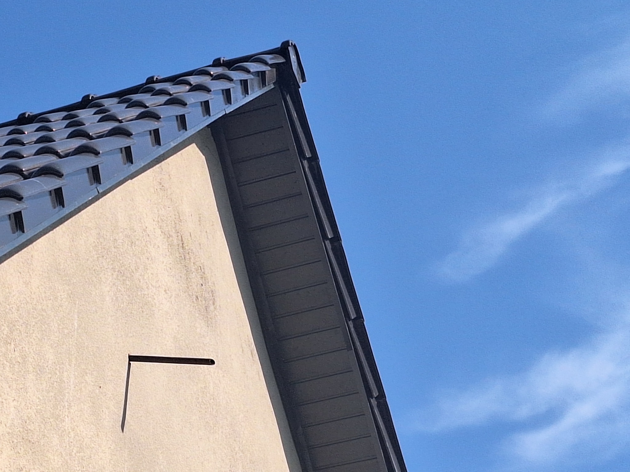 Fragment dachu z ciemnej dachówki ceramicznej, widok z dołu na tle błękitnego nieba, widoczny fragment ściany budynku z otworem wentylacyjnym.