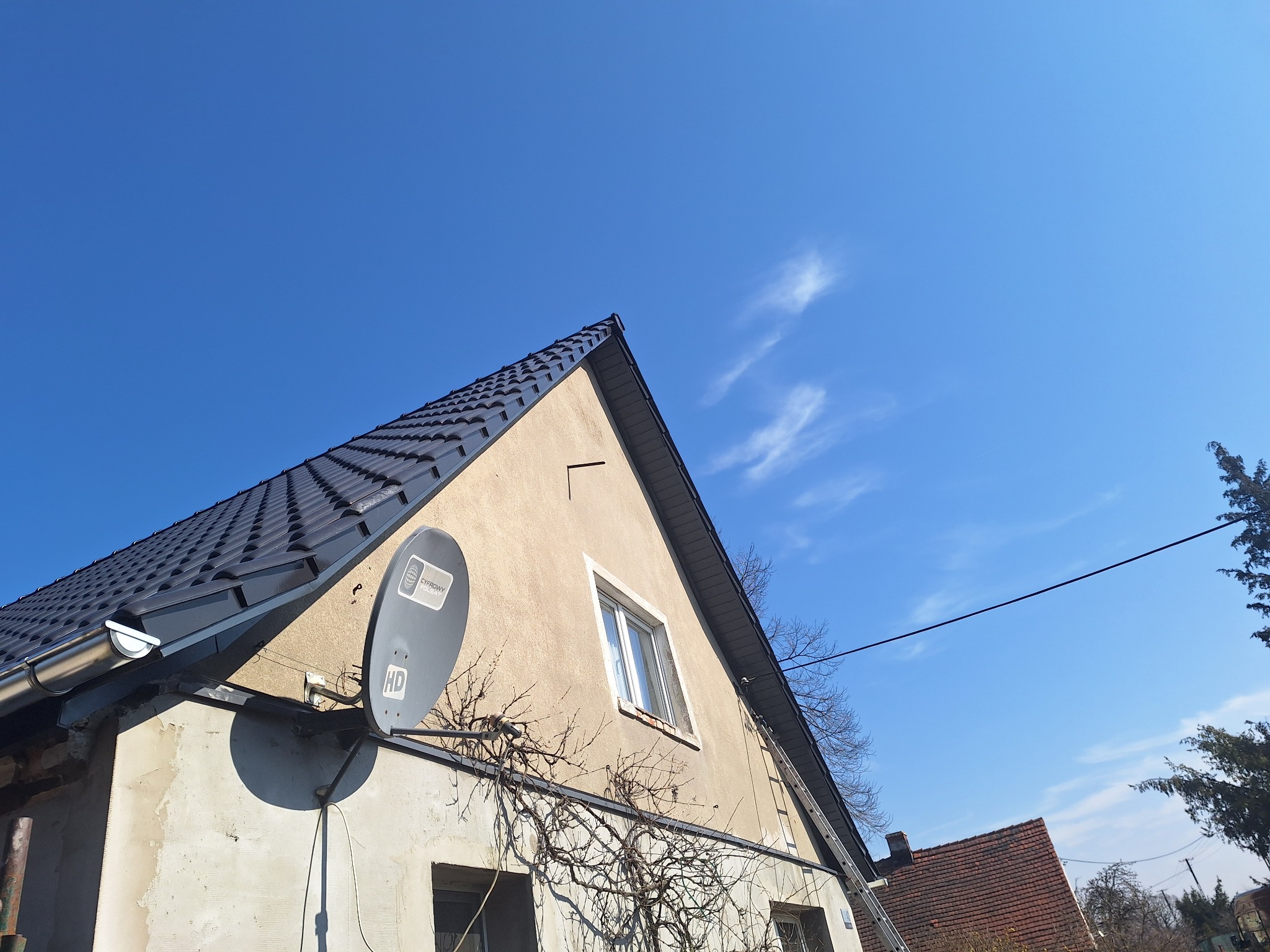 Dom z nowym, ciemnym dachem z dachówki ceramicznej, widoczna antena satelitarna i drabina oparta o ścianę. Błękitne niebo w tle.