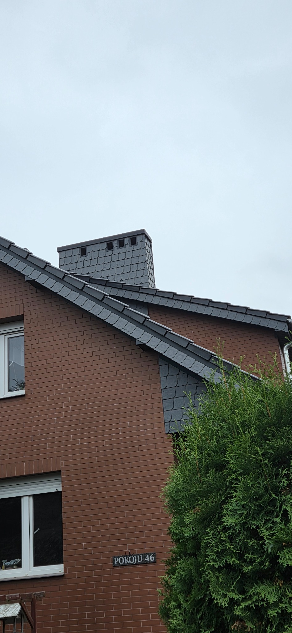 Część domu z brązowej cegły z dachem pokrytym szarą dachówką łuskową i kominem. Widoczny fragment zielonego krzewu i tabliczka z adresem.