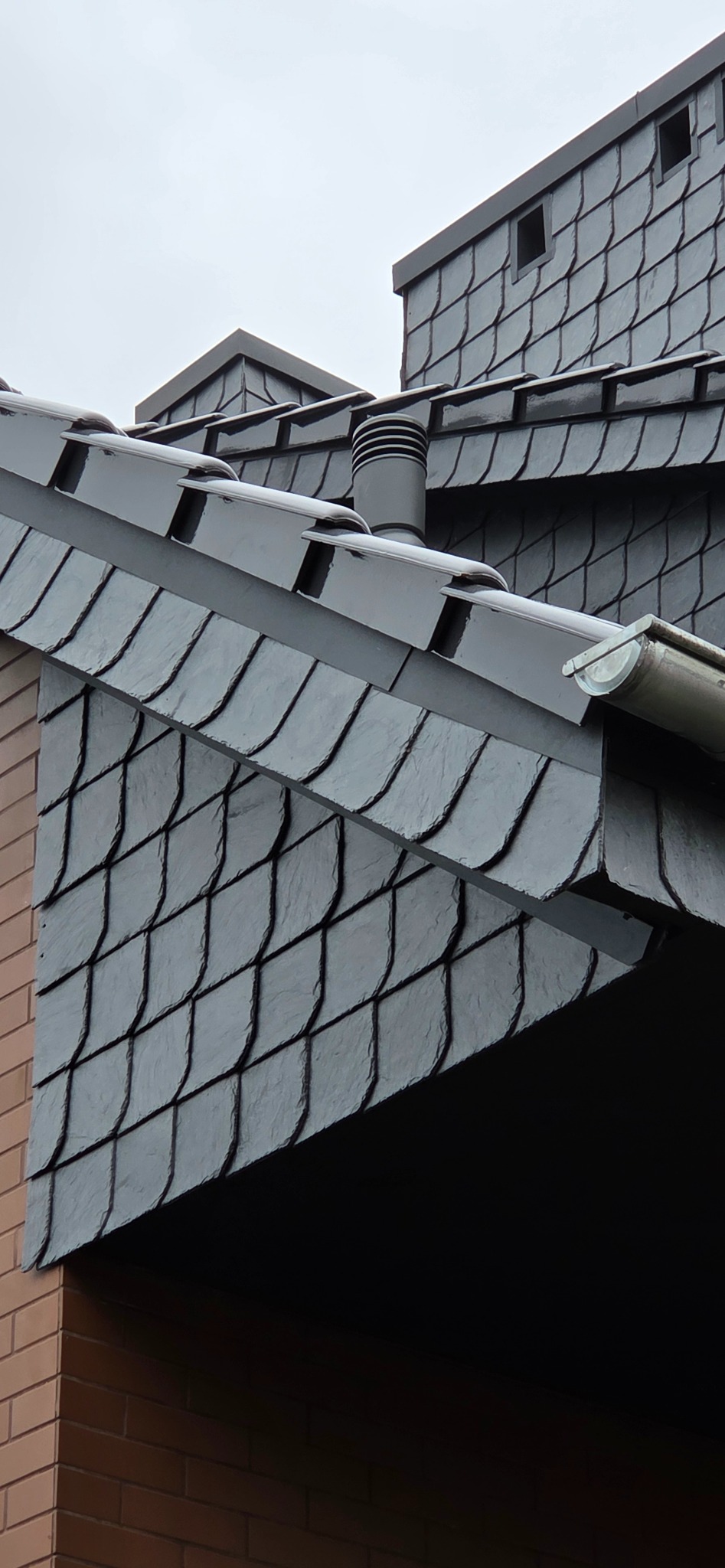 Szczegółowe ujęcie dachu krytego łupkiem z elementami wentylacji i orynnowania w kolorze grafitowym, na tle pochmurnego nieba i fragmentu ceglanej ściany.