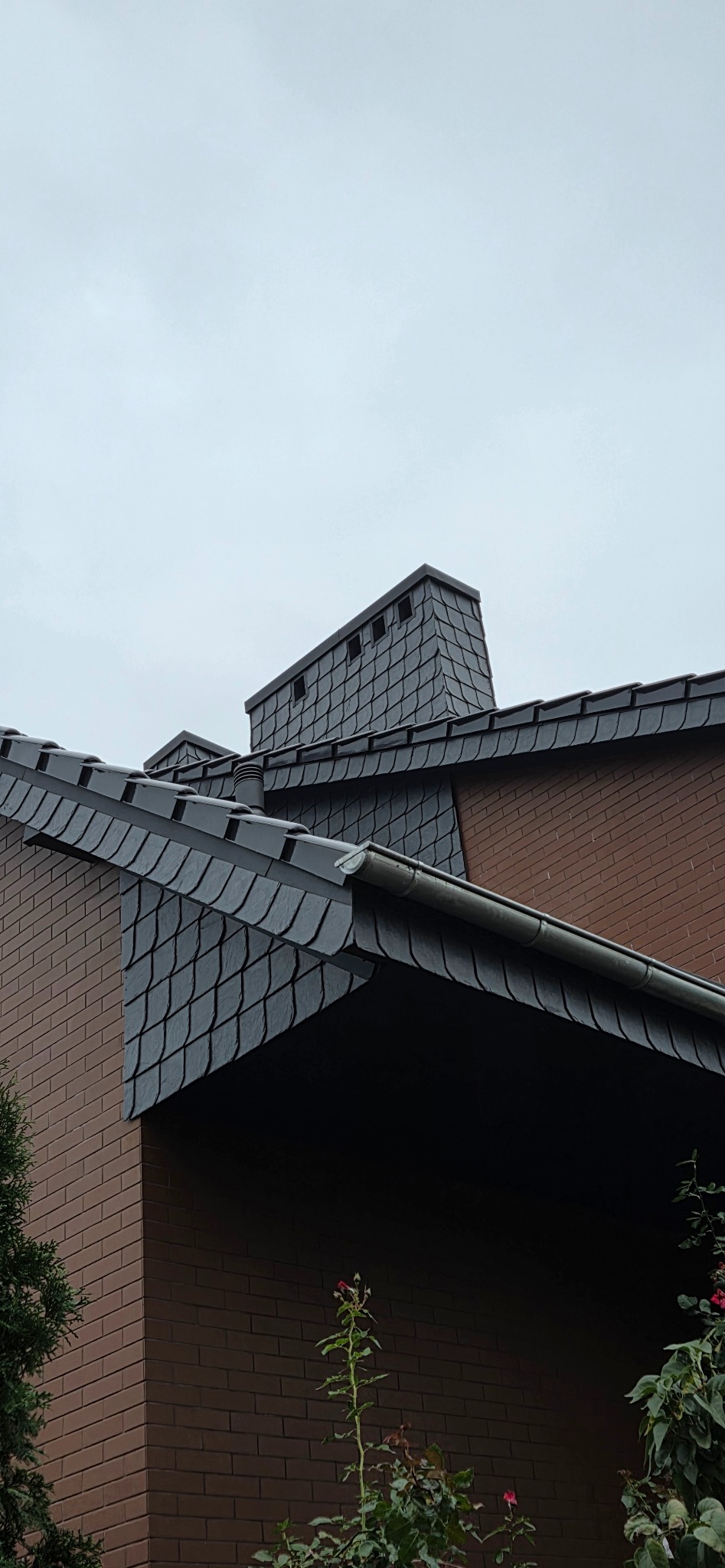 Szary dach z łupka z kominem na tle pochmurnego nieba, widoczna ceglana ściana budynku i fragment roślinności.