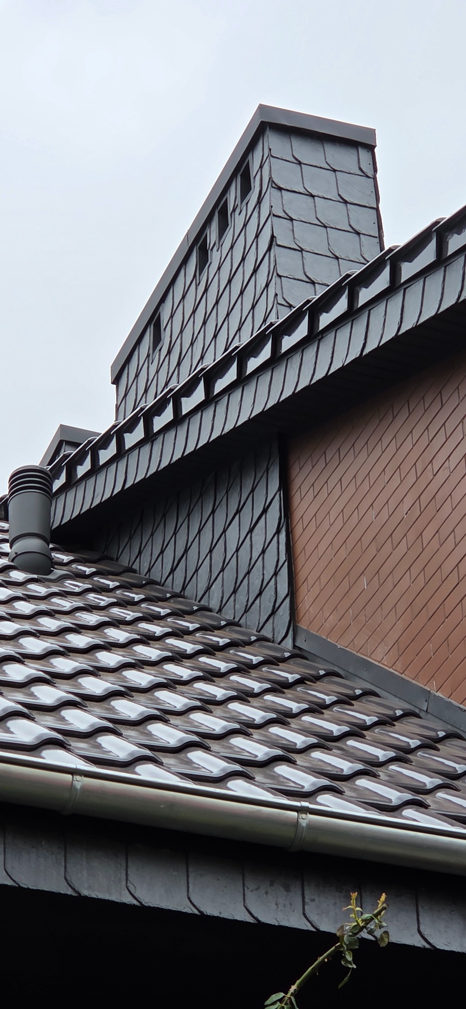 Szczegółowe ujęcie dachu krytego ciemną dachówką ceramiczną, z widocznym kominem obłożonym łupkiem i fragmentem ściany z cegły klinkierowej. Rynna w dolnej części kadru.