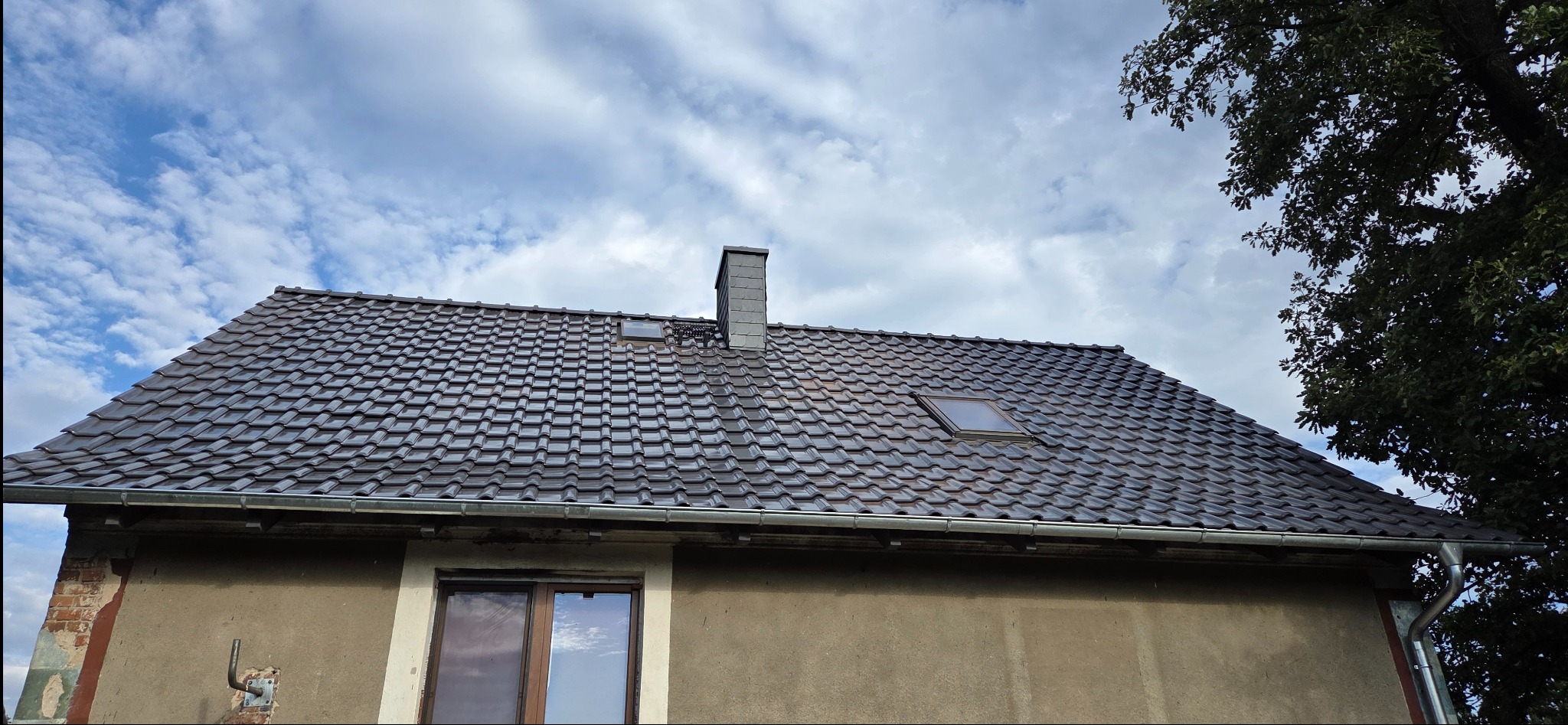 Dachówka ceramiczna w kolorze grafitowym na budynku jednorodzinnym z oknem dachowym i kominem. Widoczne rynny i fragment ściany budynku. Pochmurne niebo w tle.