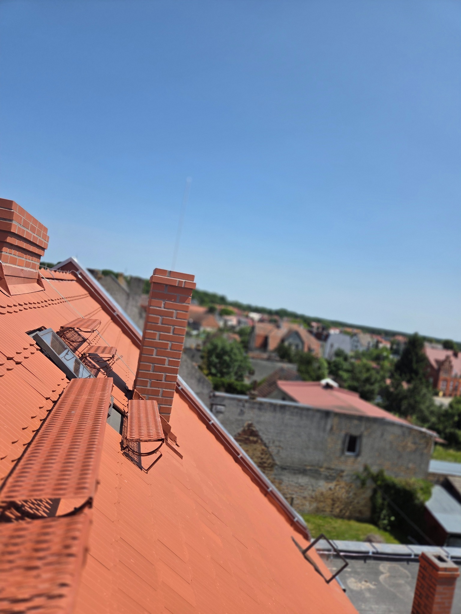 Czerwony dach z ceglanymi kominami i oknami dachowymi w słoneczny dzień. Widok na zabudowę miejską w tle, z perspektywy z góry.
