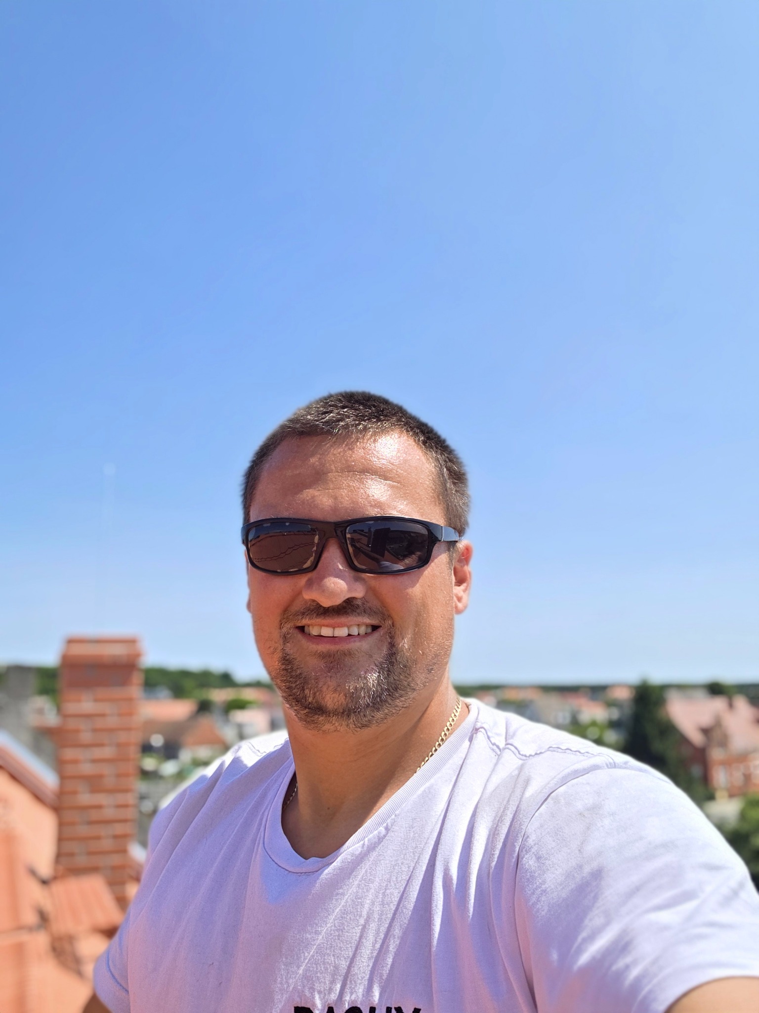 Uśmiechnięty mężczyzna w okularach przeciwsłonecznych na tle błękitnego nieba i dachu z czerwonej dachówki, widok z góry.