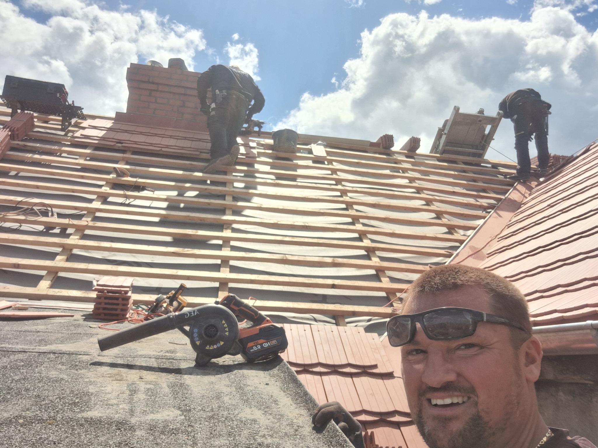 Ekipa dekarzy w trakcie krycia dachu czerwoną dachówką, widoczny komin z cegły, drewniana konstrukcja i narzędzia. Uśmiechnięty dekarz w okularach w pierwszym planie.