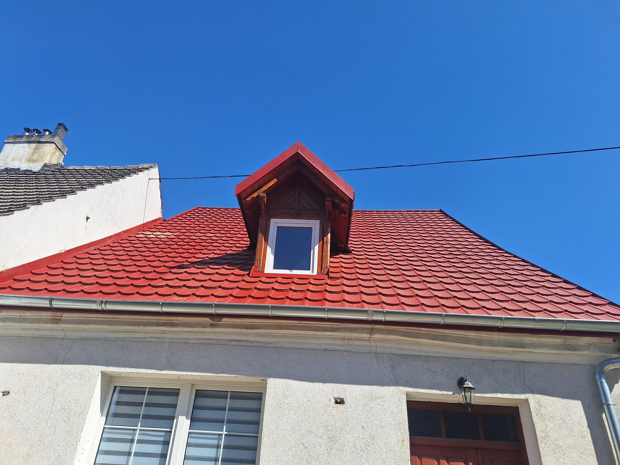 Czerwony dach z oknem dachowym i nową rynną na tle błękitnego nieba. Widoczna część budynku z oknami. Dachówka karpiówka.