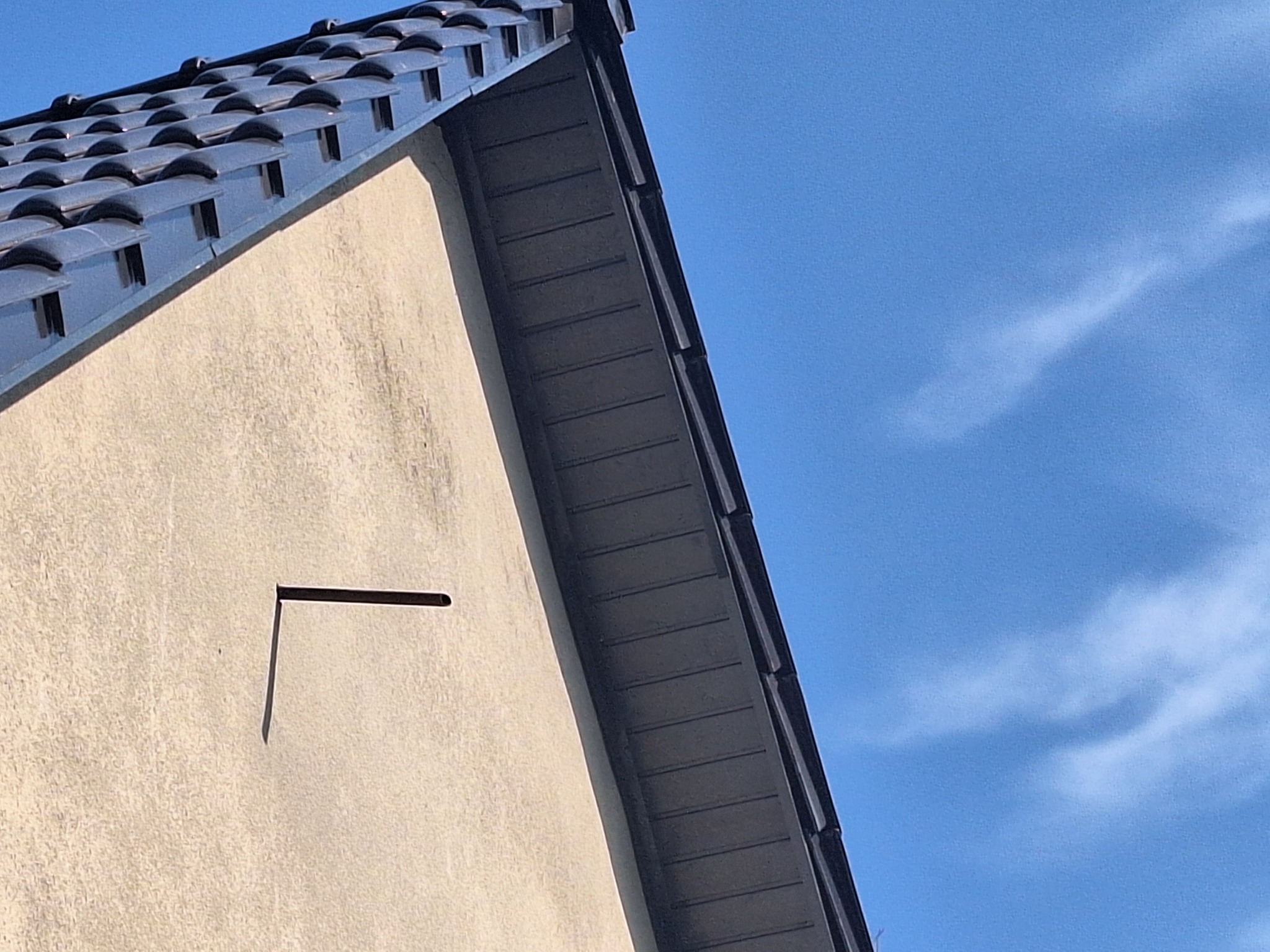 Fragment dachu z ciemną dachówką ceramiczną i fragment ściany budynku z elementem mocowania na tle błękitnego nieba.