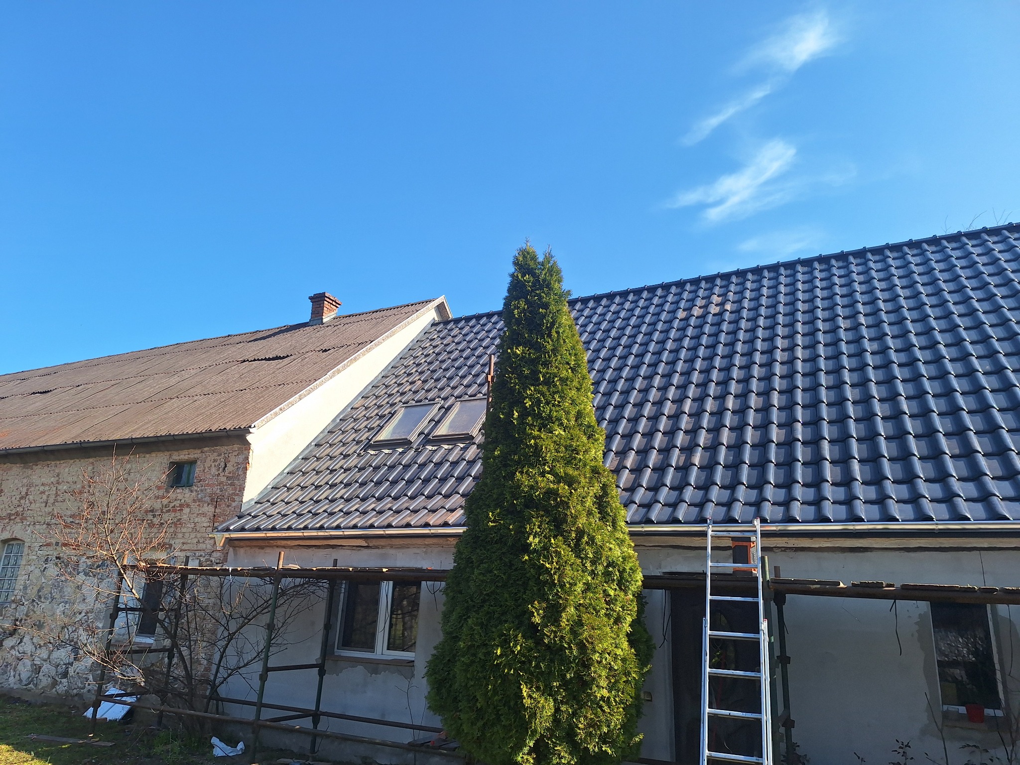 Dom z nowym, ciemnym dachem z dachówki ceramicznej, widoczna drabina oparta o ścianę. W tle stary budynek z cegły i niebieskie niebo.