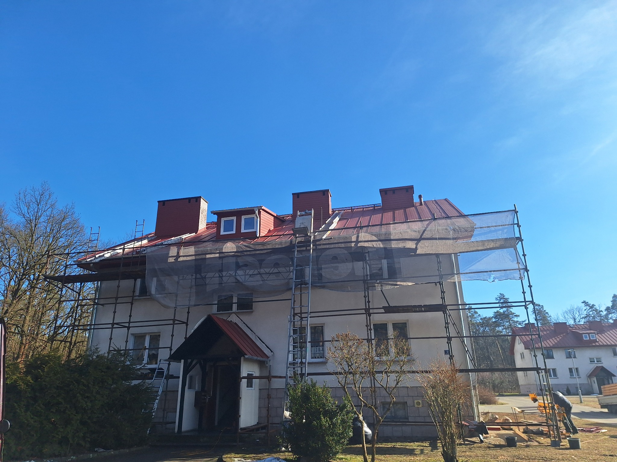 Budynek mieszkalny w rusztowaniach, przygotowany do prac na dachu z czerwonej blachy. Widoczne drabiny i zabezpieczenia. Błękitne niebo w tle.