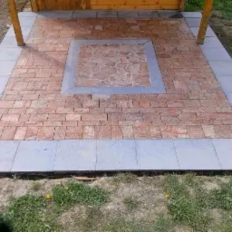 Wykończony taras z czerwono-brązowej kostki, obramowany szarymi płytami. Centralny element z mozaiki w odcieniach czerwieni. Widoczne drewniane podpory zadaszenia.