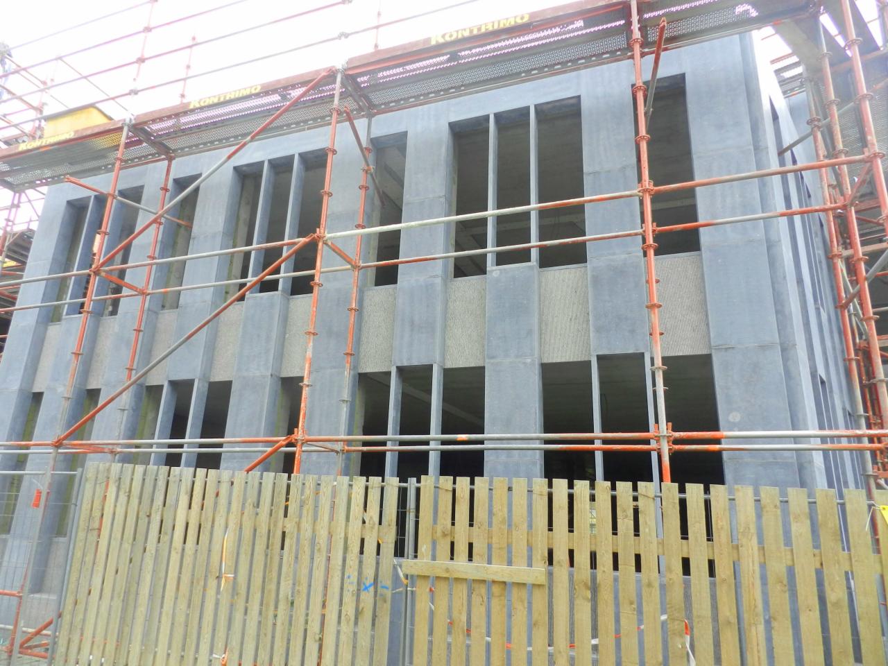 Szara fasada budynku w trakcie budowy, z widocznymi rusztowaniami i drewnianym ogrodzeniem w dolnej części kadru. Elementy kamienne w konstrukcji.