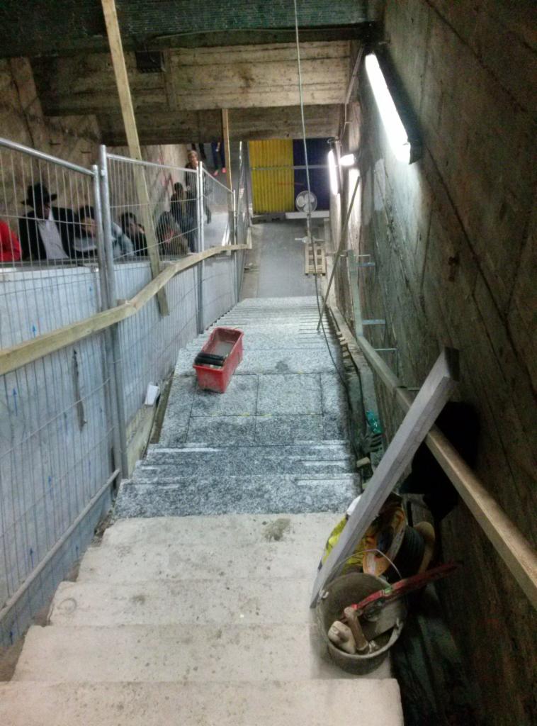 Montaż kamiennych płyt na schodach wewnątrz budynku. Widoczny kubeł z narzędziami i poziomica. Częściowo wykończone schody z szarego kamienia.