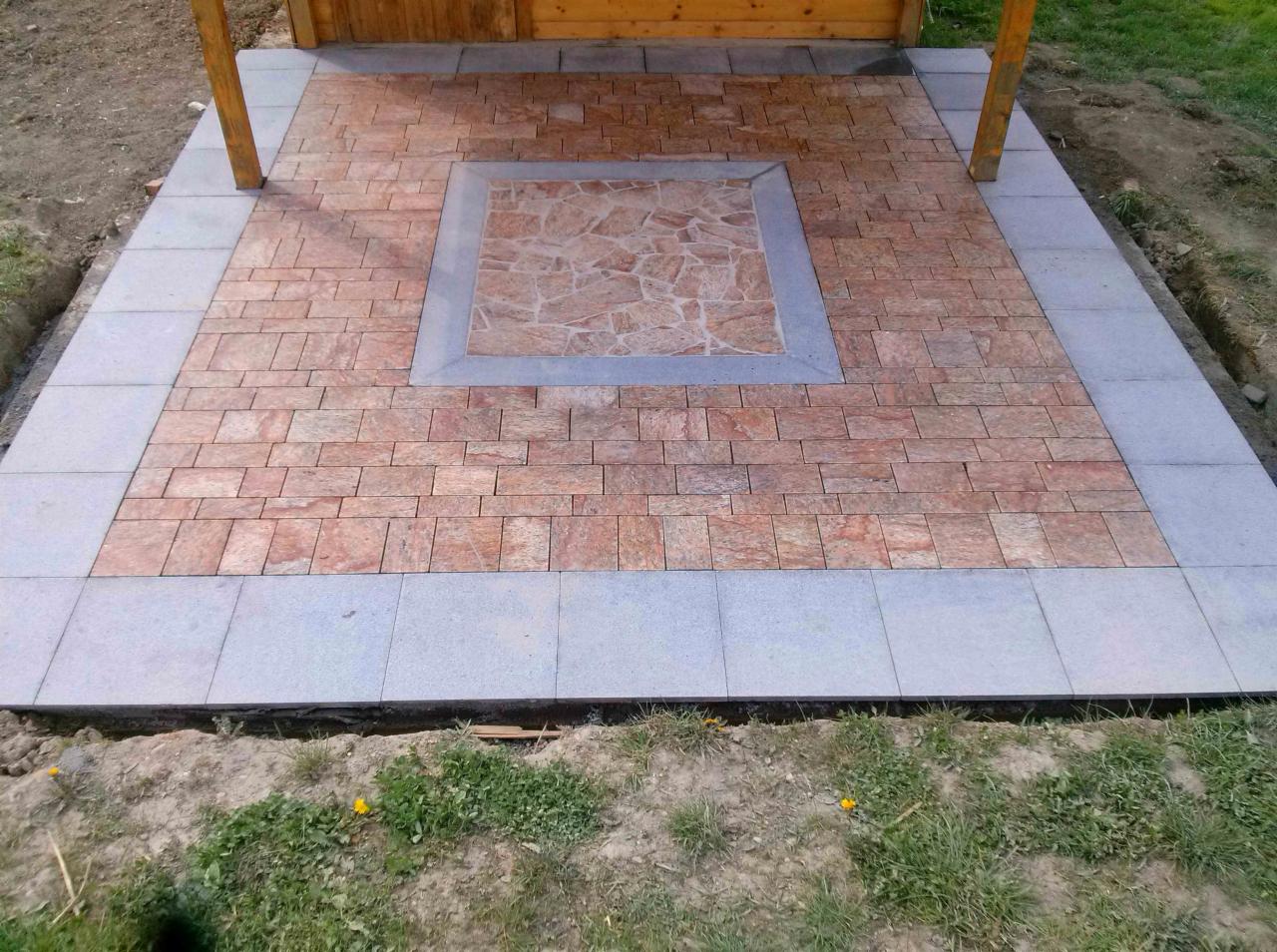 Wykończony taras z czerwono-brązowej kostki, obramowany szarymi płytami. Centralny element z mozaiki w odcieniach czerwieni. Widoczne drewniane podpory zadaszenia.