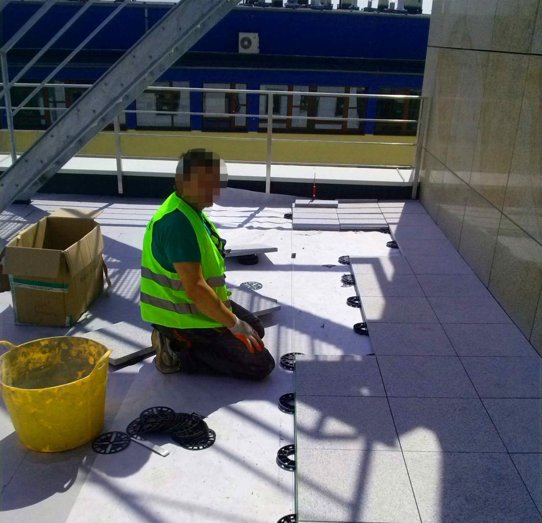 Układanie jasnych płytek na tarasie przez pracownika w odblaskowej kamizelce. Widoczne podkładki dystansowe i wiadro z zaprawą.