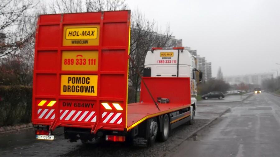 Czerwona laweta Hol-Max z Wrocławia, zaparkowana przy drodze. Widoczny numer telefonu i napis 'Pomoc Drogowa'.