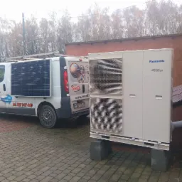 Biała pompa ciepła Panasonic Inverter na betonowych podstawach, obok zaparkowany biały samochód dostawczy z panelami fotowoltaicznymi na dachu, z logo i numerem telefonu firmy na boku.