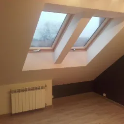Poddasze z dwoma oknami dachowymi, grzejnikiem i wykończonymi ścianami w odcieniach beżu i brązu, podłoga laminowana imitująca drewno.
