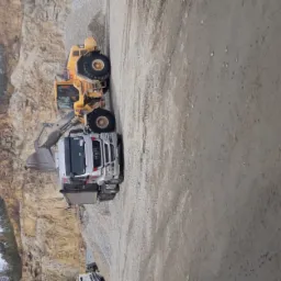 Żółta ładowarka nabiera materiał do srebrnej ciężarówki MAN na tle skalistej ściany kamieniołomu.