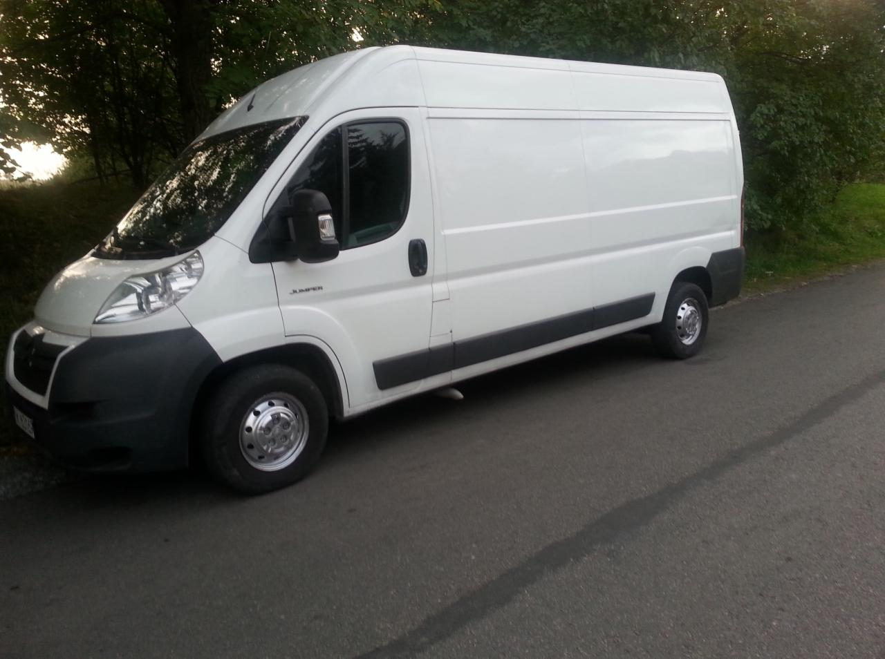 Biały bus marki Citroen Jumper, zaparkowany na asfalcie, z widocznymi chromowanymi kołpakami i ciemnymi szybami. Tło stanowią drzewa i krzewy.