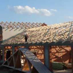 Wznoszenie więźby dachowej na murowanym budynku. Widoczny pracownik na konstrukcji. Elementy drewniane, cegły, beton. Niebo z chmurami w tle.