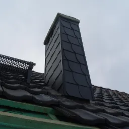 Dach z ciemnej dachówki ceramicznej z obróbką komina łupkiem. Widoczny fragment barierek śniegowych. Ujęcie z dołu na szare niebo.