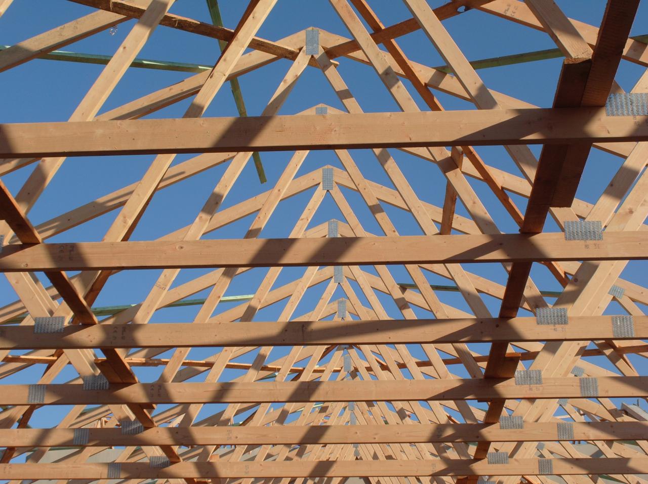 Drewniana konstrukcja dachu z widocznymi elementami złącznymi na tle błękitnego nieba. Widok z dołu, ukazujący geometrię i regularność konstrukcji.