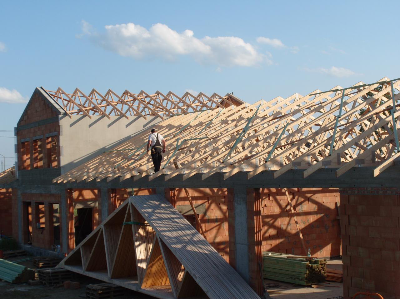 Wznoszenie więźby dachowej na murowanym budynku. Widoczny pracownik na konstrukcji. Elementy drewniane, cegły, beton. Niebo z chmurami w tle.
