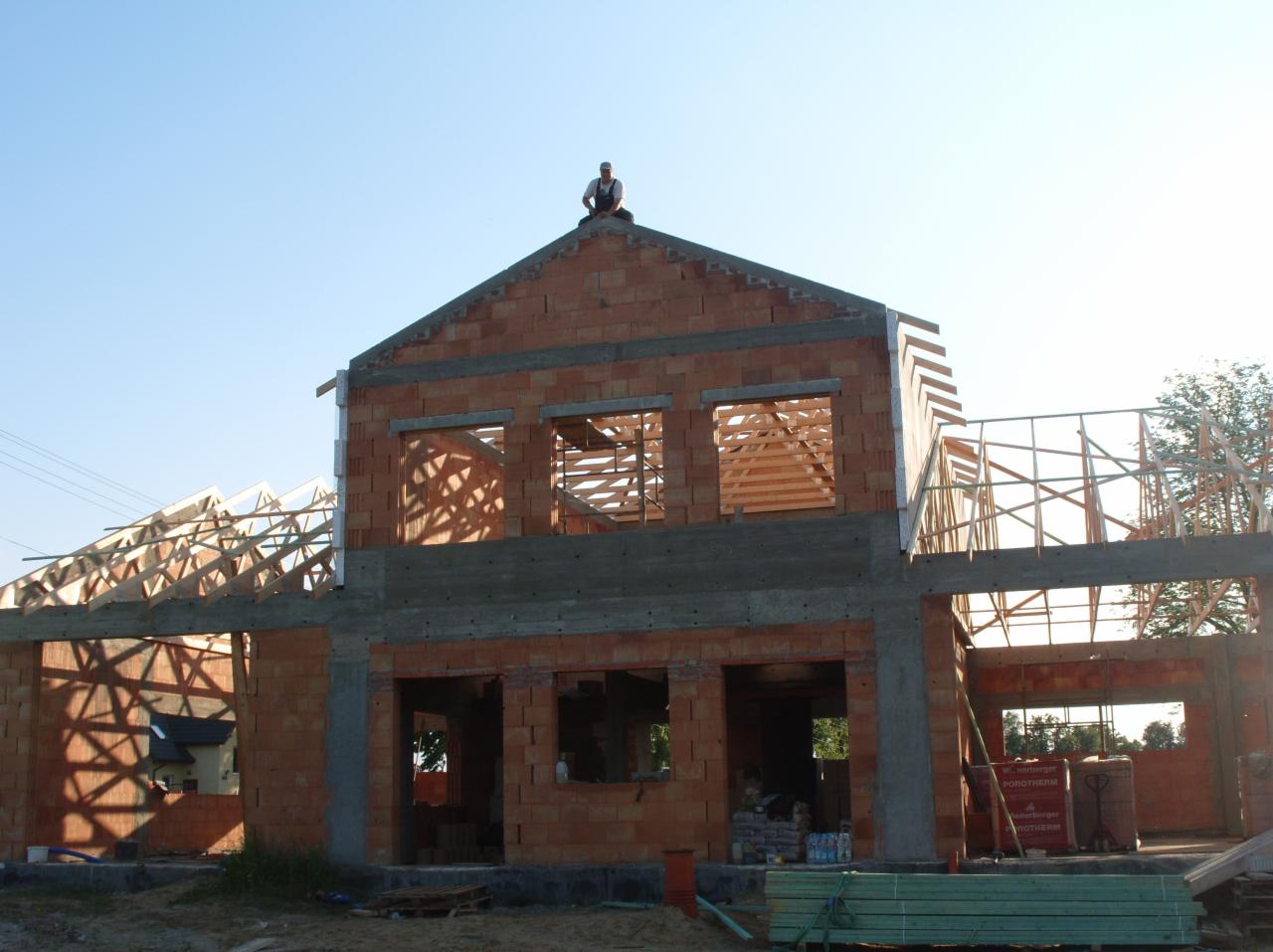 Budowa domu z czerwonej cegły Porotherm, widoczne zbrojenia i drewniana konstrukcja dachu. Na szczycie siedzi osoba. Materiały budowlane u podstawy.
