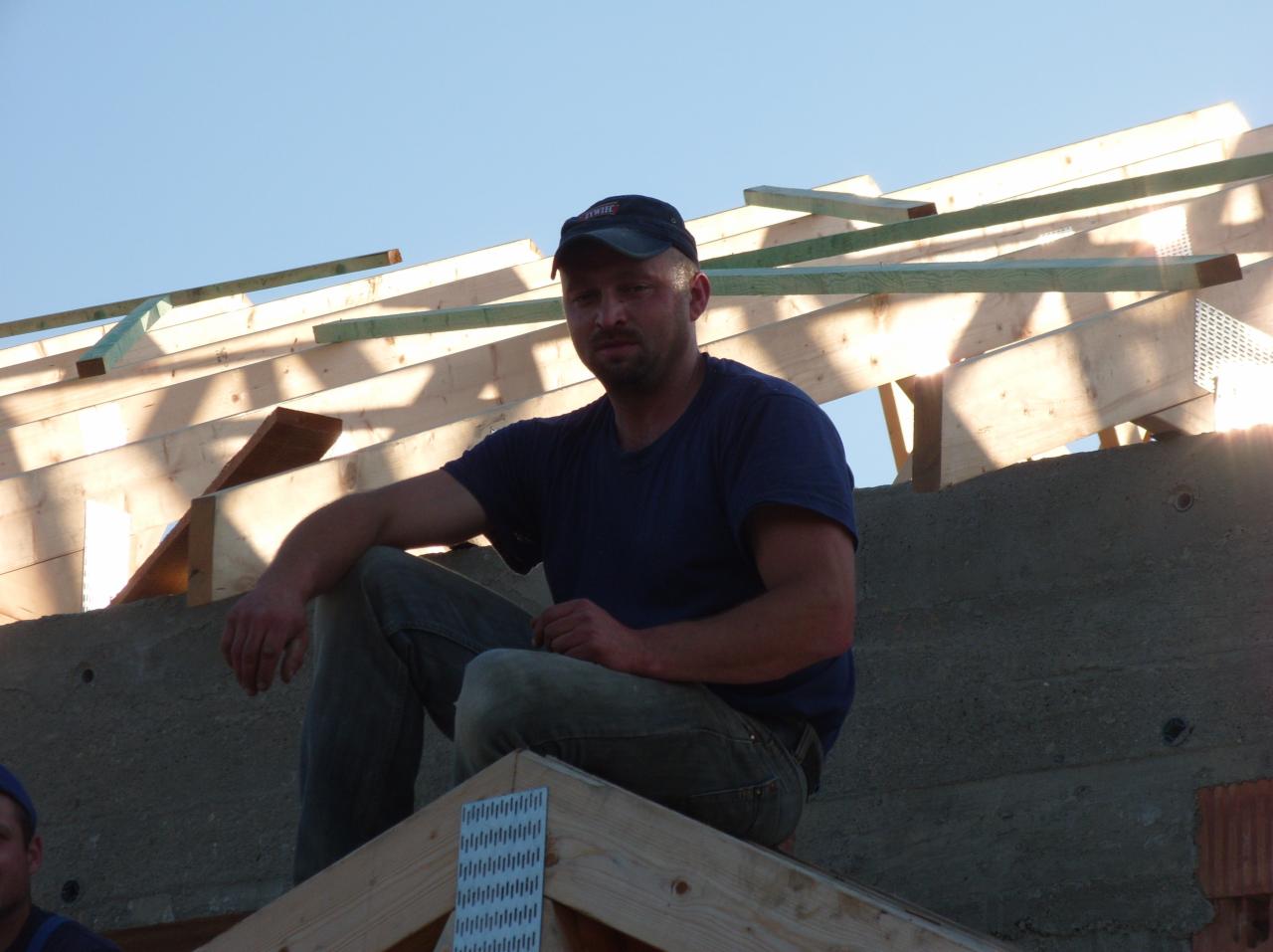 Pracownik w czapce siedzi na drewnianej konstrukcji dachu w trakcie budowy, z widocznymi elementami muru i niebem w tle. Widok na konstrukcję dachu w budowie.