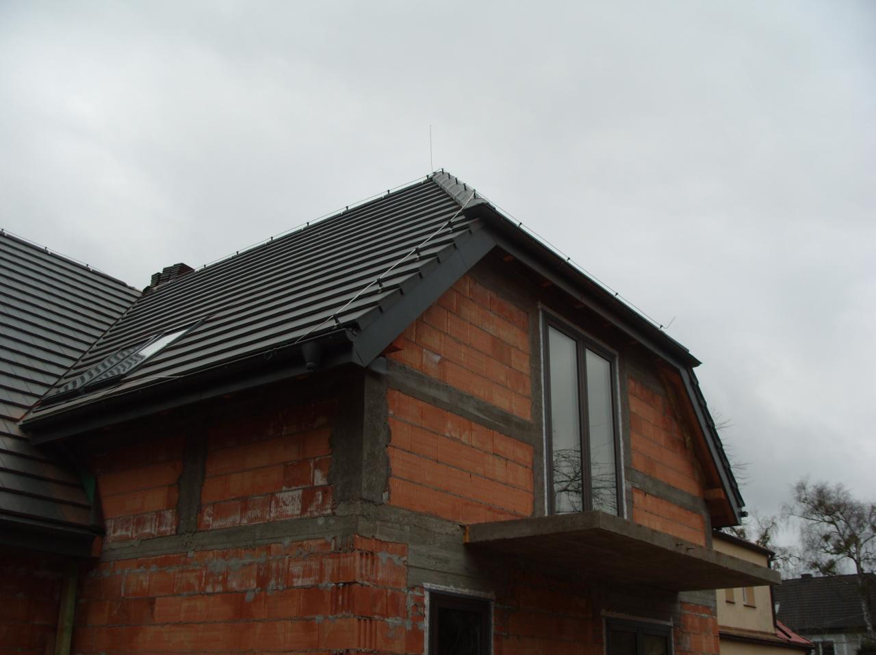 Dom w trakcie budowy z ciemną dachówką i oknem balkonowym. Widoczny stan surowy, cegła i beton. Dach z orynnowaniem i odgromnikiem.