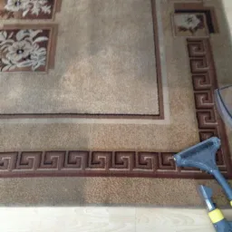 Czyszczenie beżowego dywanu z wzorem za pomocą dyszy parowej. Widoczne fragmenty parkietu i narzędzia czyszczącego.