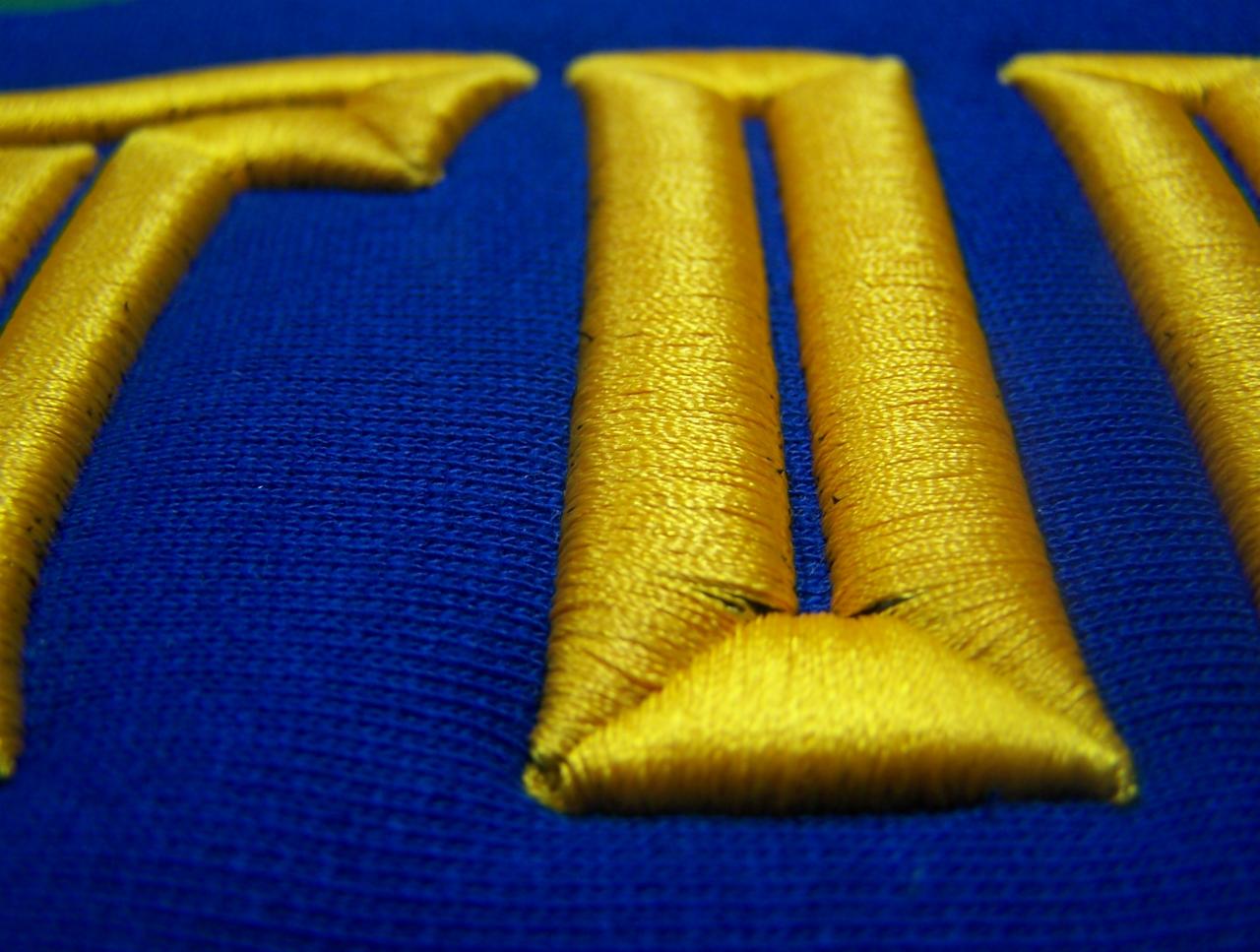 Detale złotego haftu na niebieskim materiale, zbliżenie na fragment napisu.