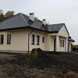 Dom jednorodzinny z beżową elewacją, czarną dachówką i kolumnami przy wejściu, widoczny z boku na tle pochmurnego nieba i drzew bez liści, z samochodem typu pickup zaparkowanym obok.