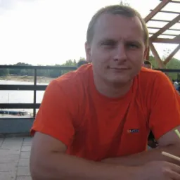 Mężczyzna w pomarańczowej koszulce siedzi przy stoliku na zewnątrz, z widokiem na jezioro i plażę w tle. Widoczne słomki w napoju i fragment zadaszenia.