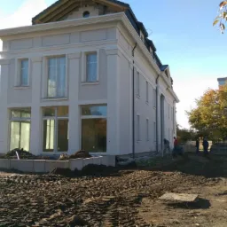 Jasna elewacja dwukondygnacyjnego domu z dużymi oknami, widoczni pracownicy, teren w trakcie prac ziemnych, elementy architektury ogrodowej.