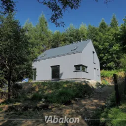 Nowoczesny, szary dom modułowy z płaskim dachem i oknami w ciemnych ramach, usytuowany na wzniesieniu z widokiem na las.