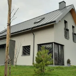 Nowoczesny dom jednorodzinny z szarą blachodachówką i elewacją w kolorze gołębim, z dużymi oknami osłoniętymi żaluzjami zewnętrznymi, na pierwszym planie mała choinka.